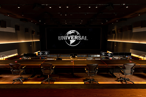 A dark editing suite featuring a long desk with multiple monitors and a large screen displaying the Universal logo.