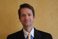 A man with brown hair wearing a dark suit, white shirt, and yellow striped tie, smiling against a yellow wall.
