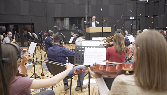 Musicians with instruments and sheet music perform in a studio guided by a conductor at a podium.