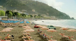 A sandy beach with blue and pink umbrellas, parked cars, and a large green mountain in the background.
