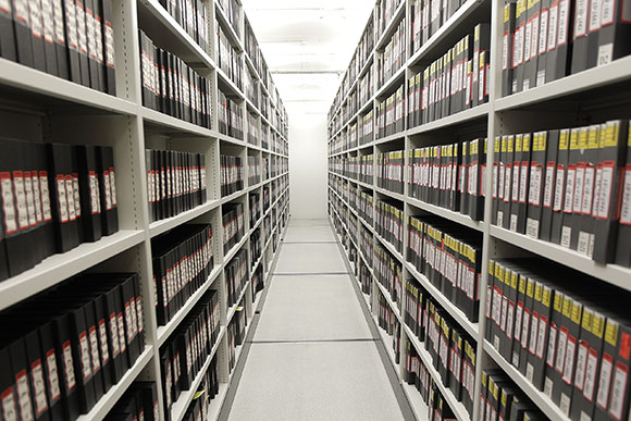 Rows of black binders with red labels line white shelves in a long, narrow archive aisle.