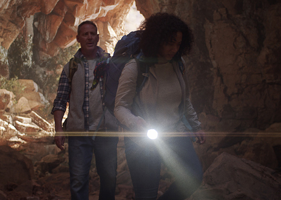 A man and a woman with backpacks walk through a dark cave using a flashlight.