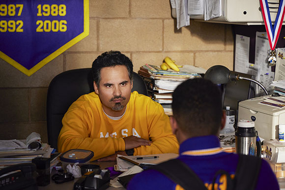 A man in a yellow sweatshirt sits at a cluttered desk, looking toward a person in a blue jacket.