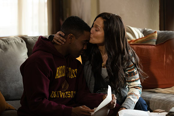 A woman leans in to kiss a young man's forehead while he sits on a couch holding a paper.