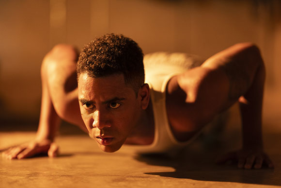 A man in a white tank top performs a push-up on a floor in warm, dim lighting.