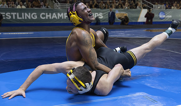 Two male wrestlers in singlets grappling on a blue mat during a wrestling match.