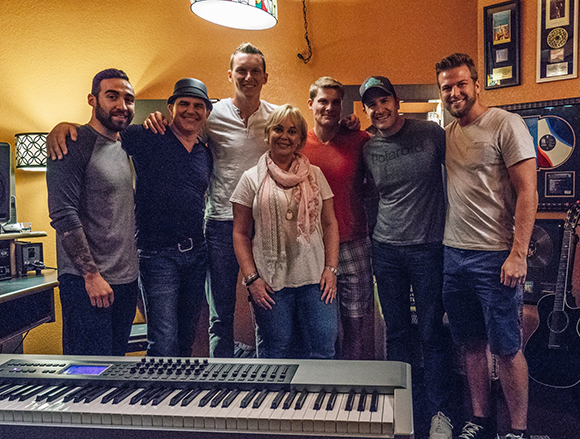 Seven people stand in a group behind a digital keyboard in a room with music memorabilia.