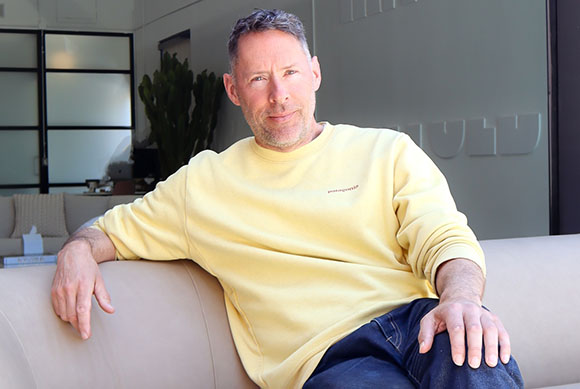 A man with short hair and light stubble wearing a yellow sweatshirt sits on a beige couch.