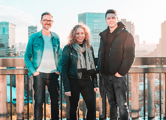 Three people stand smiling on a rooftop with a cityscape in the background.