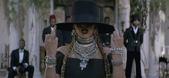 A woman in a wide-brimmed black hat and ornate silver jewelry gestures with her middle fingers.