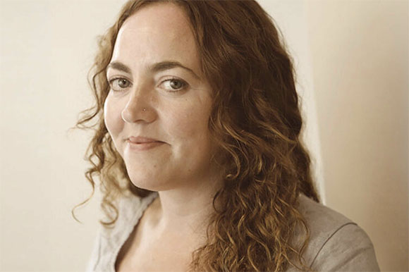 A woman with long, wavy brown hair looks toward the camera with a slight smile.