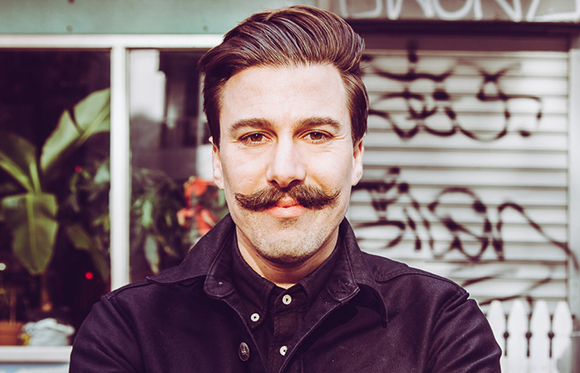 A man with dark hair and a prominent handlebar mustache wearing a black button-down shirt.
