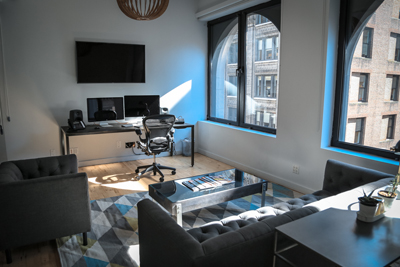 A bright home office featuring a desk with dual monitors, a black office chair, and large arched windows.