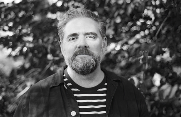 Black and white portrait of a man with a beard wearing a striped shirt against a leafy background.