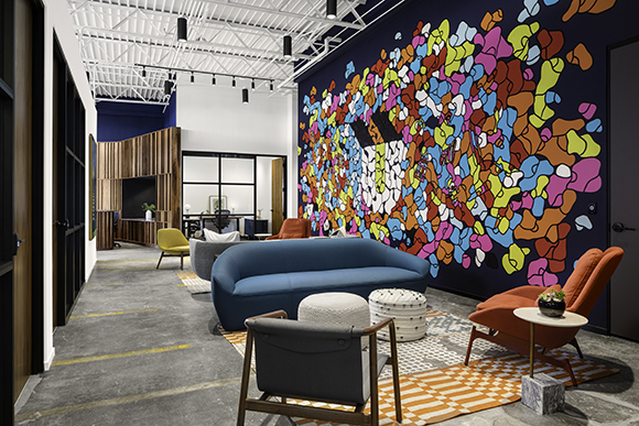 A modern lounge area featuring a blue sofa, various armchairs, and a large colorful abstract mural on a dark wall.