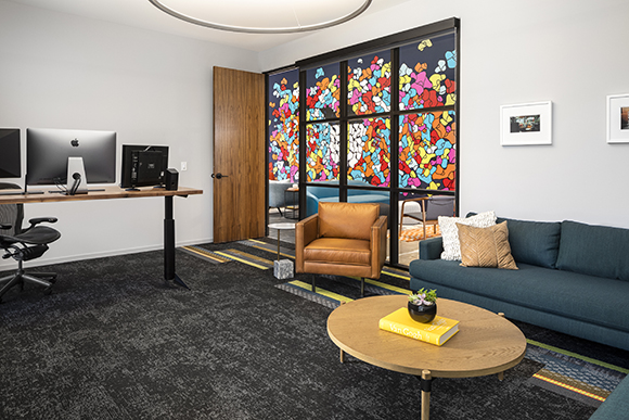 Modern office with a wooden desk, computer, leather armchair, blue sofa, and a colorful glass partition.