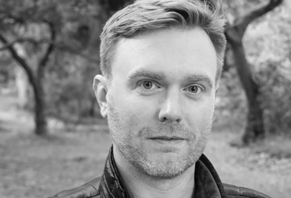 Black and white close-up portrait of a man with short hair looking directly at the camera in a wooded area.