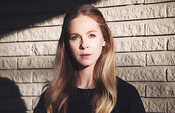 A woman with long blonde hair looks forward against a sunlit brick wall.