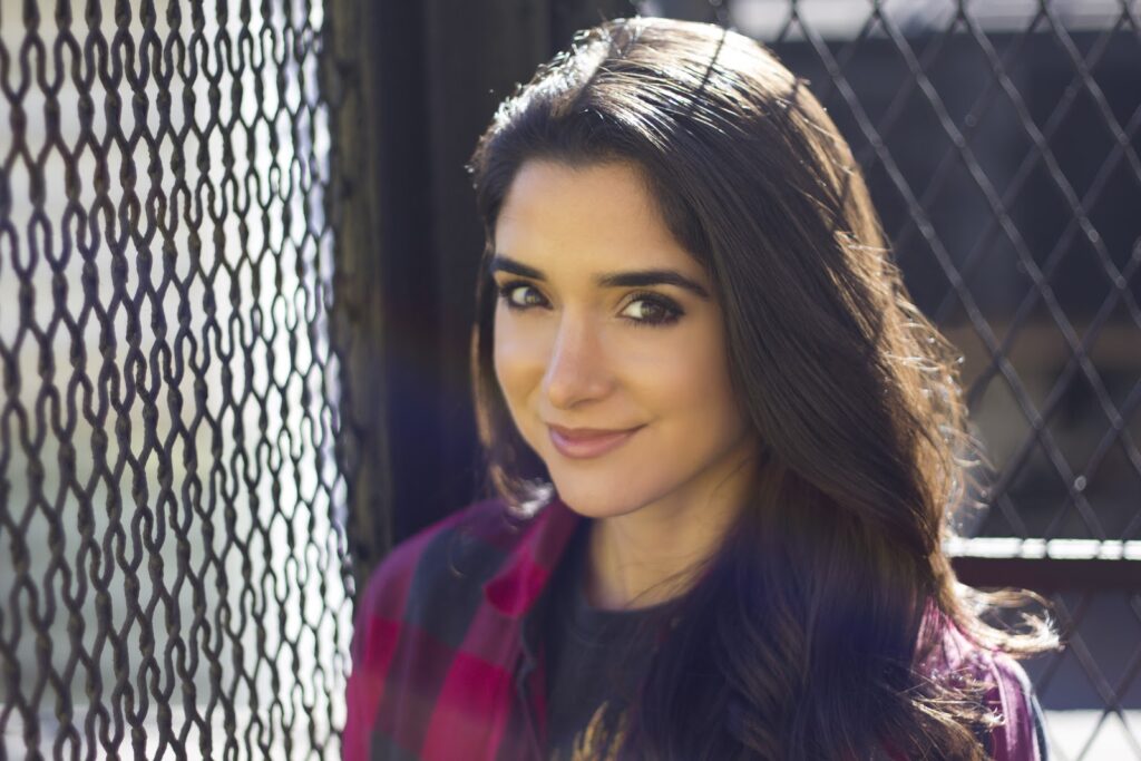 A woman with long dark hair smiles while standing in front of a chain-link fence.