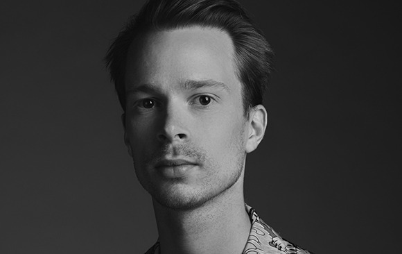 Black and white close-up portrait of a young man looking directly at the camera against a dark background.
