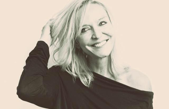A smiling blonde woman with her hand in her hair, wearing a dark off-the-shoulder top.