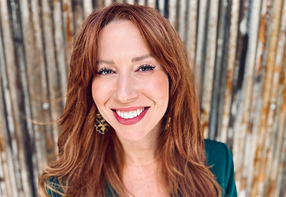 A smiling woman with long reddish-brown hair and large patterned earrings against a wooden background.