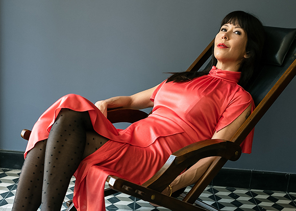 A woman with dark hair and red lipstick reclines in a dark wooden lounge chair wearing a red silk dress.