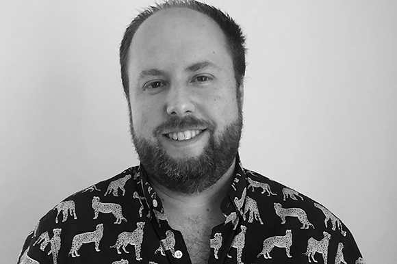 Black and white portrait of a smiling man with a beard wearing a dark shirt with a leopard pattern.