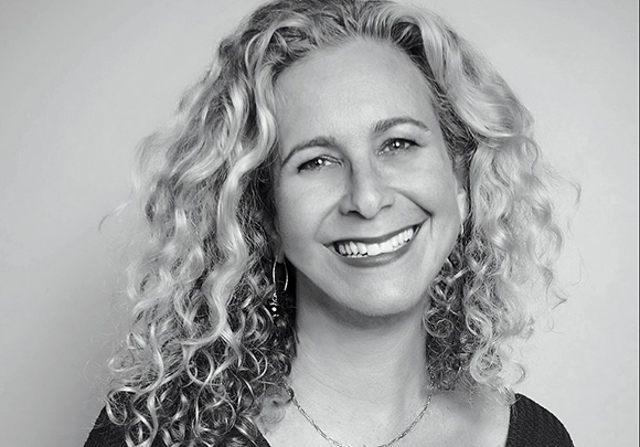 A smiling woman with long, light-colored curly hair looking toward the camera.