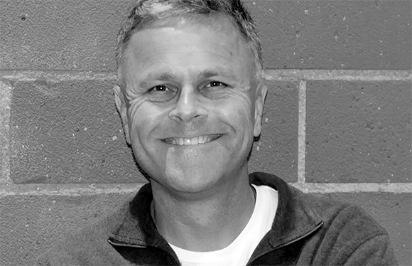 A smiling middle-aged man with short hair wearing a zip-up jacket over a white t-shirt.