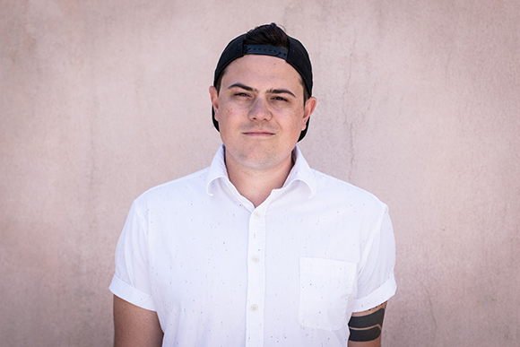 A man wearing a white button-down shirt and a black baseball cap facing forward against a pink wall.