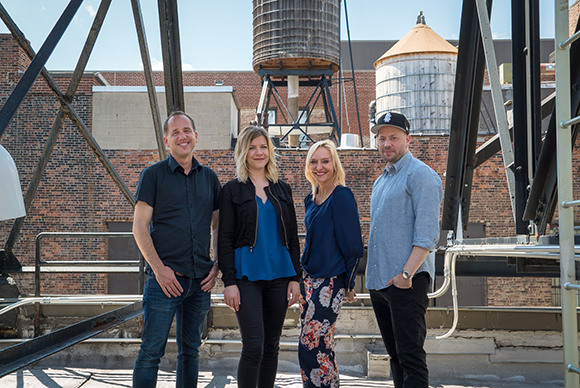 Four smiling adults standing on a rooftop with industrial water towers and brick buildings behind them.