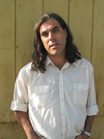 A man with long dark hair and a white button-down shirt stands against a yellow wooden wall.