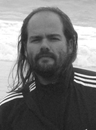 A man with long hair and a beard wearing a dark athletic jacket with white stripes on the shoulders.