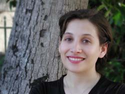A smiling woman with dark hair leaning against a large tree trunk outdoors.