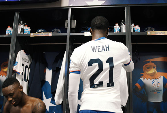 A soccer player wearing a white jersey with WEAH 21 on the back stands in a locker room.
