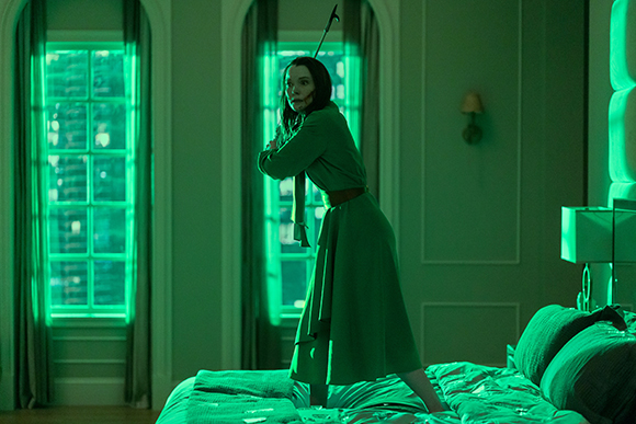 A woman in a green dress stands on a bed in a room bathed in green light.