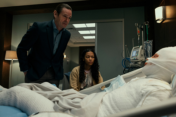 A man in a suit and a young woman look down at a patient wrapped in white bandages in a hospital bed.