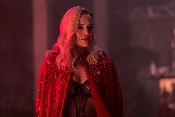 A blonde woman wearing a red lace cape and black lace bodysuit stands in dim, red-toned lighting.