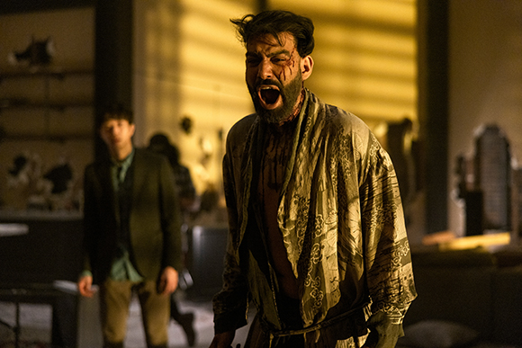 A man with blood on his face and chest screams intensely while wearing an open patterned robe.
