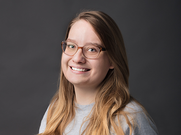 A smiling woman with long blonde hair and brown-rimmed glasses against a dark gray background.