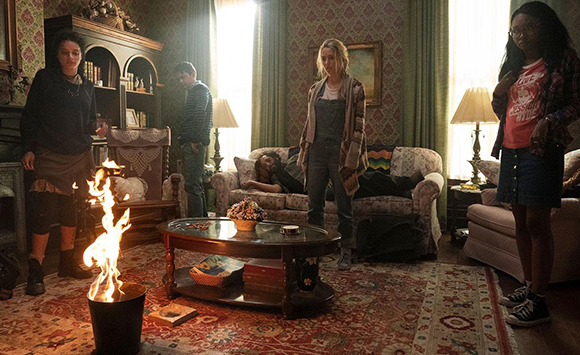 Four people stand in a cluttered living room around a small fire burning in a black bucket on a patterned rug.