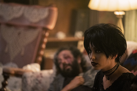 A woman with dark hair and heavy makeup looks sideways in a dimly lit room with a man behind her.