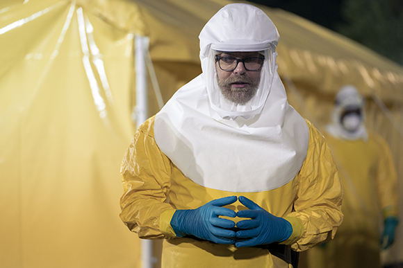 A person wearing a yellow hazmat suit, white hood, and blue gloves stands in front of a yellow tent.