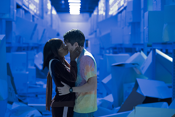 A young man and woman embrace and kiss in a large warehouse filled with boxes under blue lighting.
