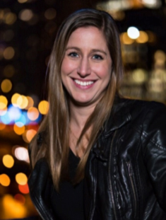 A smiling woman with long blonde hair wearing a black leather jacket against a blurred city background.