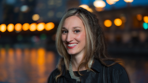A woman with long blonde and dark dreadlocked hair smiles against a bokeh city background at night.