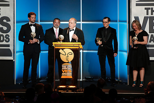 Five people in formal attire stand on a stage holding awards in front of a blue paneled backdrop.