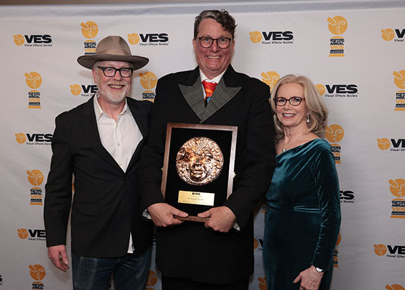 Three people stand together in front of a VES Awards backdrop, with the center man holding a gold award.