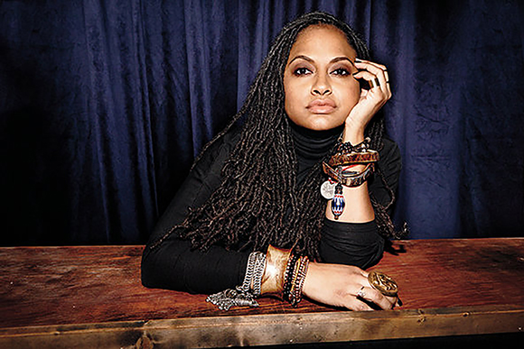 A woman with long dreadlocks and many bracelets leans her head on her hand against a dark blue curtain.